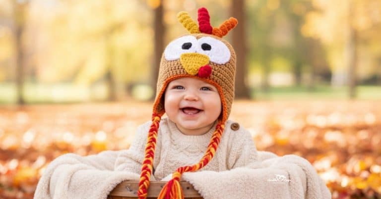Baby wearing a crochet turkey hat with braided tassels while sitting in a basket outdoors surrounded by colorful fall leaves