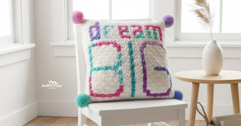 &ldquo;Dream Big&rdquo; crochet pillow with colorful pom poms displayed on a white chair in a bright neutral room.