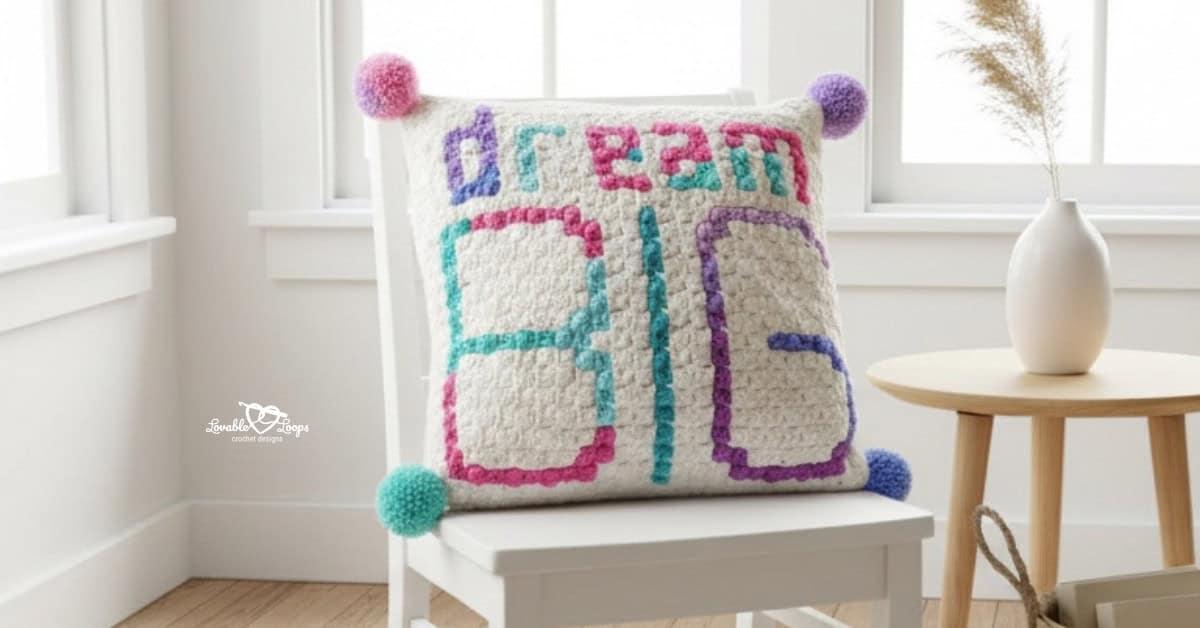 “Dream Big” crochet pillow with colorful pom poms displayed on a white chair in a bright neutral room.
