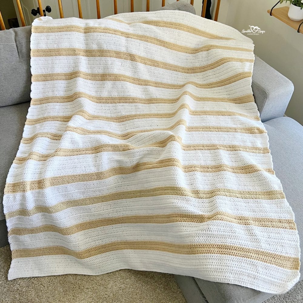 Neutral striped double crochet blanket draped across a gray sofa, featuring soft white and tan stripes with textured back loop detail
