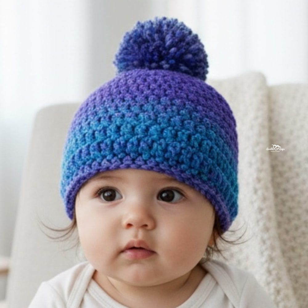 Baby wearing a purple and blue crochet beanie with a matching pom-pom while sitting in a cozy chair.