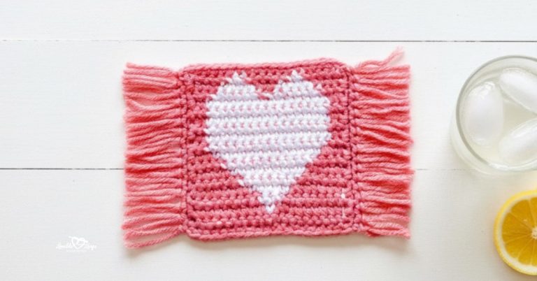 Top-down photo of a pink crochet heart coaster with fringe on both sides and a white heart design, styled on a whitewashed table beside a glass of lemonade and a lemon slice.