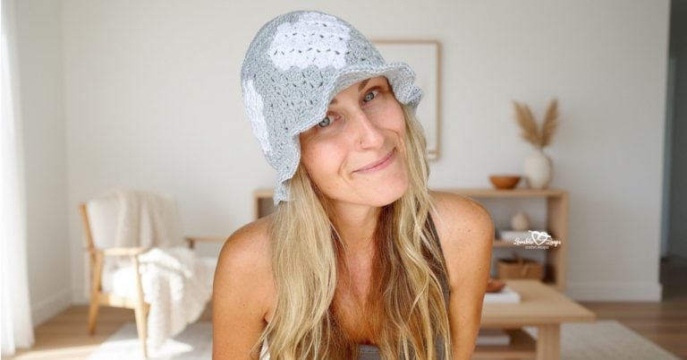 Woman wearing a gray and white crochet heart bucket hat, photographed in a bright neutral living room with soft natural light.
