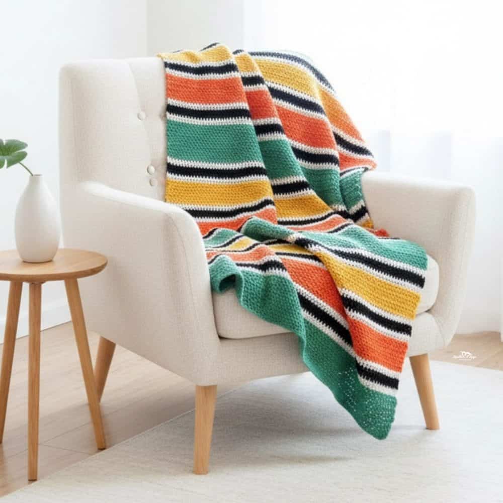 Striped crochet afghan draped over a cream armchair in a bright neutral room, featuring bold green, orange, mustard, black, and white horizontal stripes.