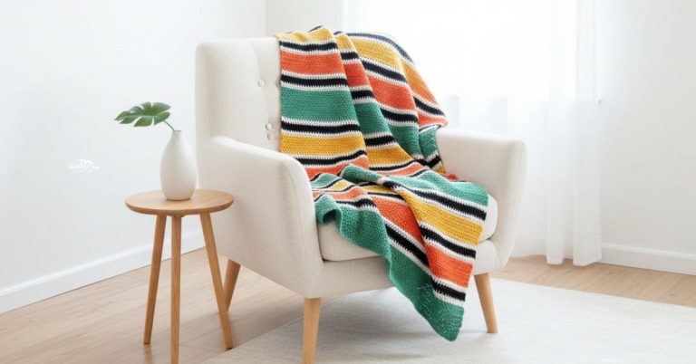 Crochet striped blanket with green, orange, yellow, black, and cream rows draped over a cream armchair in a bright room.