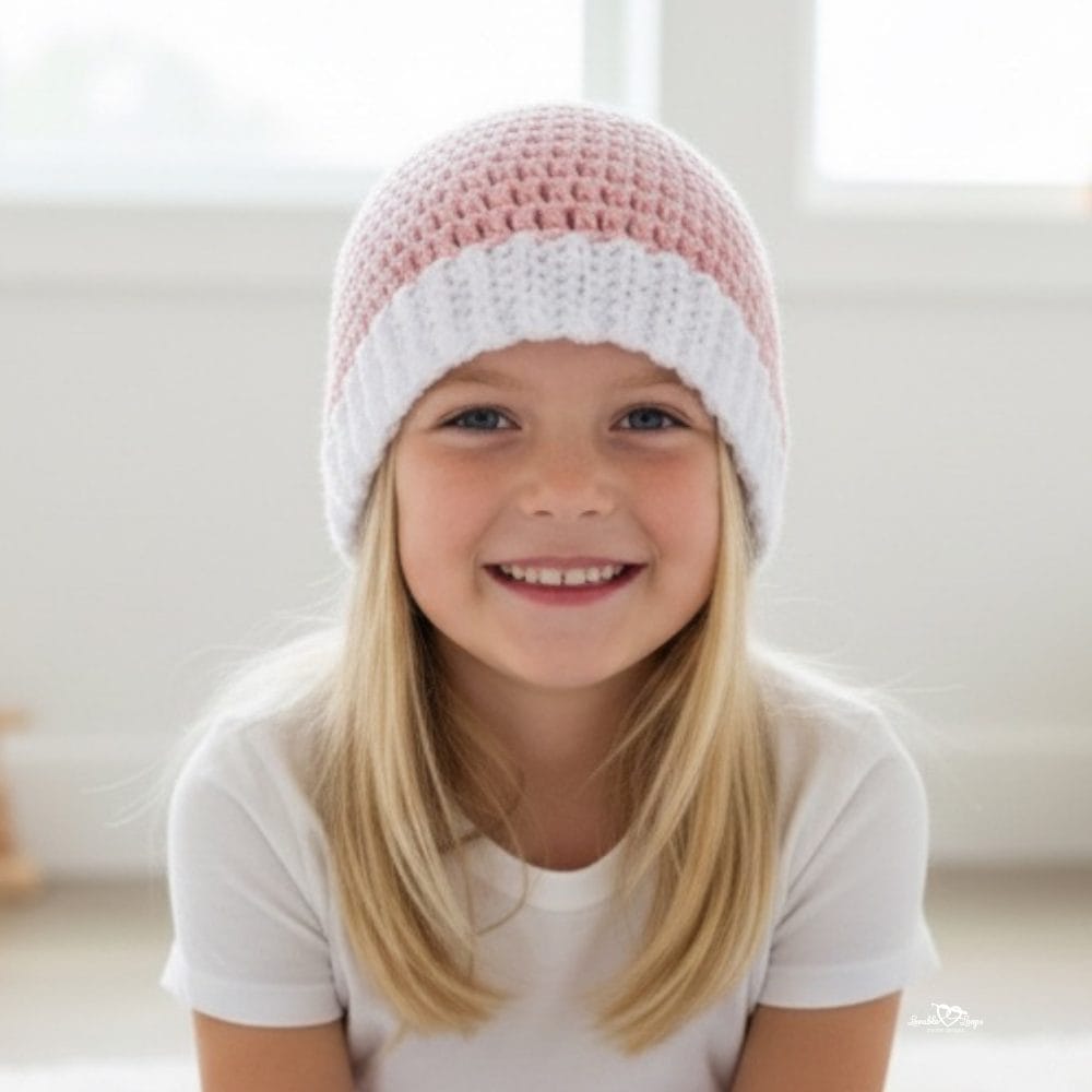 Smiling girl wearing a pink and white crochet beanie in a bright neutral room.