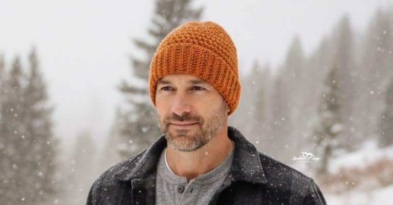 Man wearing an orange crochet beanie, standing outdoors in a snowy winter landscape with pine trees in the background.