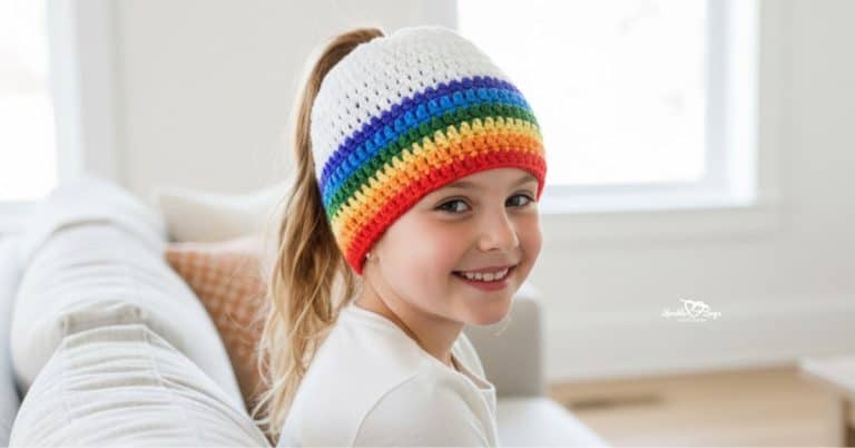 Child wearing a white rainbow-striped crochet ponytail hat with a high ponytail, sitting on a couch in a bright cozy living room.