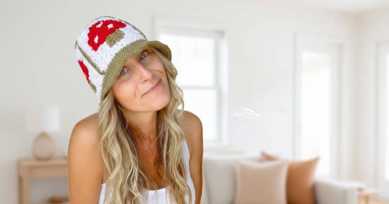 Woman wearing a mushroom crochet bucket hat with red and tan details, photographed in a bright neutral living room with soft natural light.