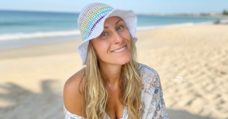 Woman wearing a white crochet rainbow bucket hat, photographed on a sunny beach with ocean in the background.