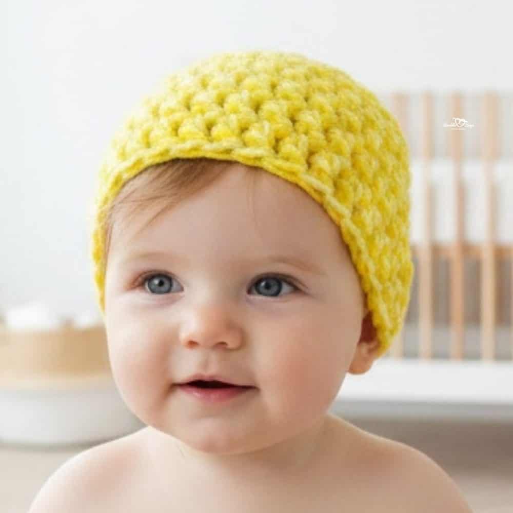 Baby wearing a bright yellow crochet beanie in a soft neutral nursery.