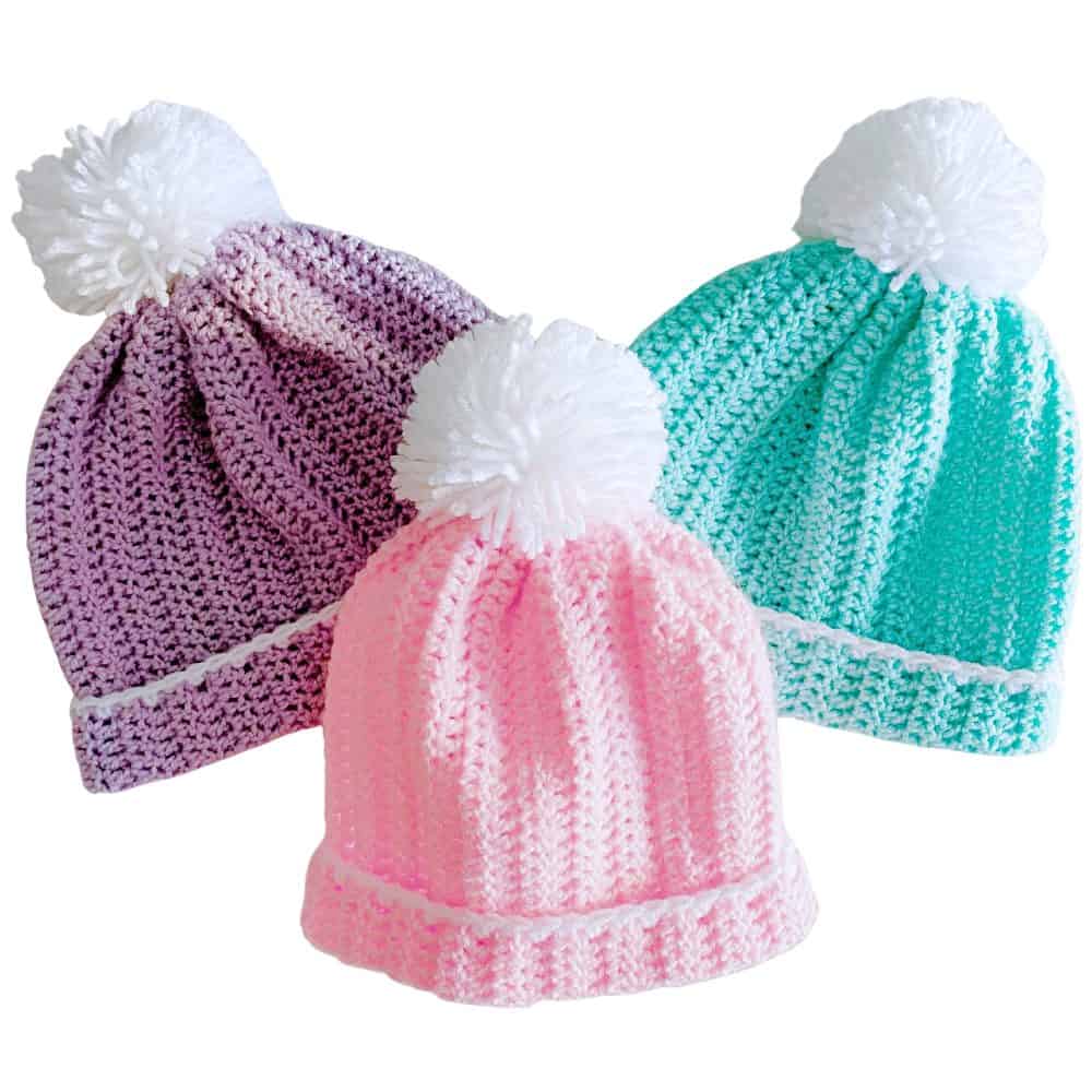 Crochet slouchy hat pattern shown in three sizes, including pink, purple, and aqua beanies with folded brims and white pom poms.