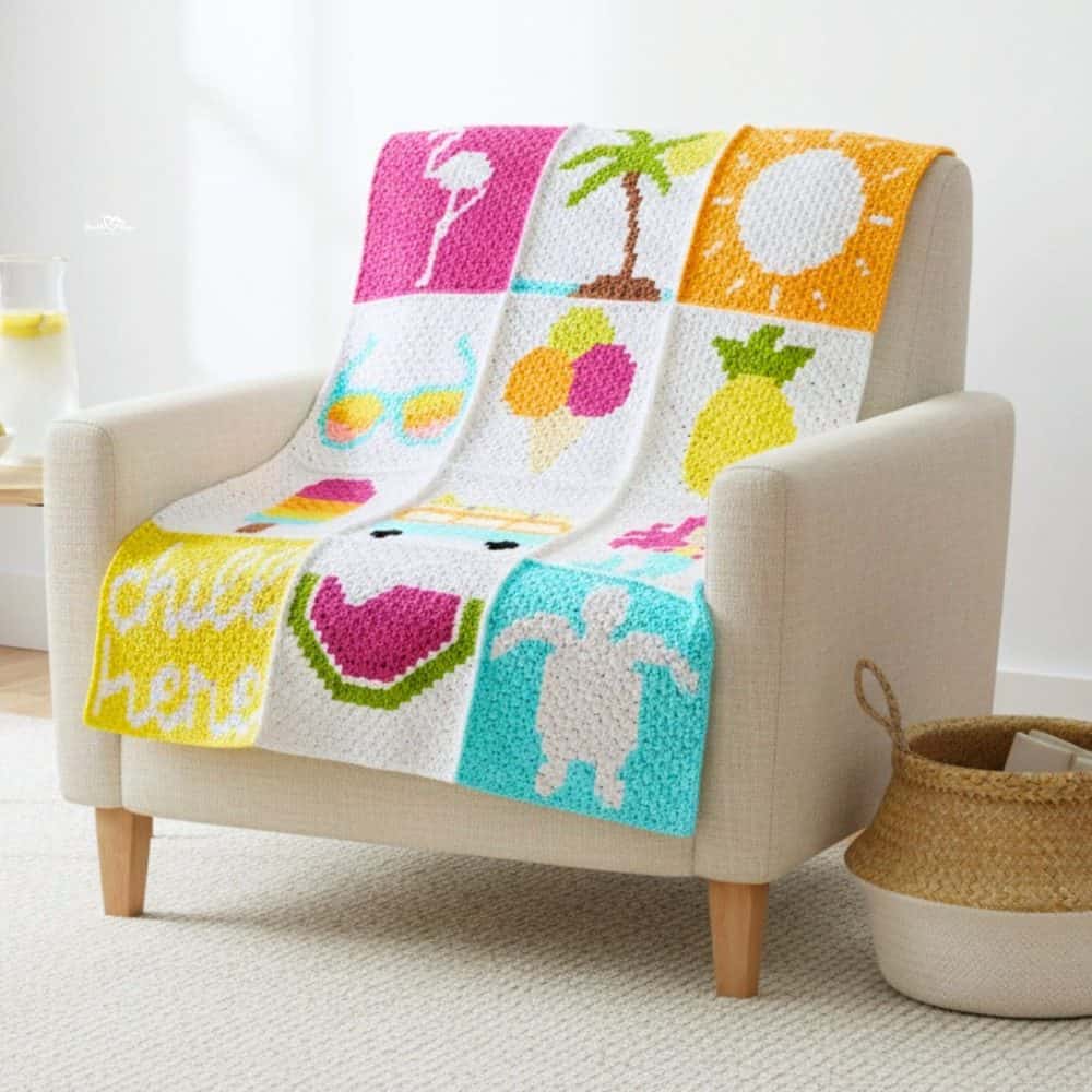 Colorful C2C summer blanket draped over a cream armchair in a bright living room, featuring summer-themed squares with a flamingo float, palm tree, sun, sunglasses, ice cream, pineapple, watermelon, sea turtle, and &ldquo;chill here&rdquo; text.