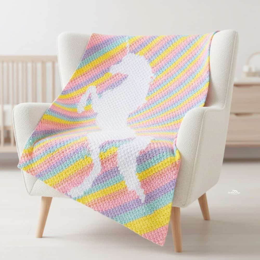 C2C unicorn crochet blanket draped over a cream armchair, featuring a white unicorn on pastel rainbow stripes.