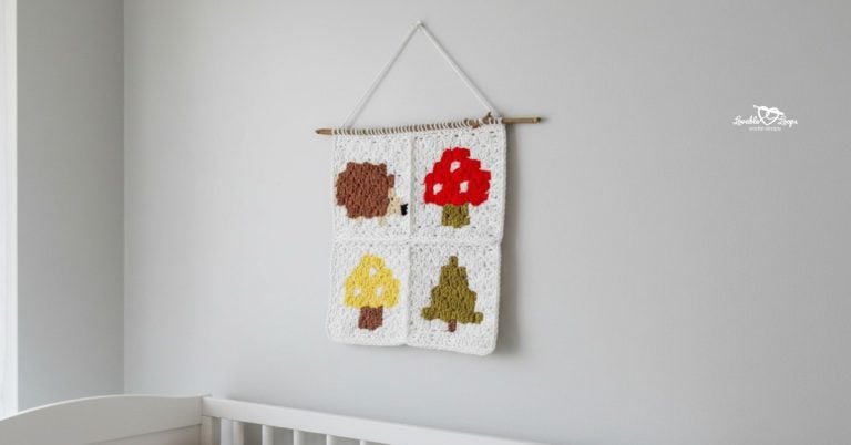 Crochet woodland wall hanging hanging above a crib in a nursery, featuring four white granny squares with a hedgehog, red mushroom, yellow mushroom, and tree.