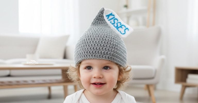 Toddler wearing a gray pointed crochet Hershey Kiss hat with a white “KISSES” tag in a bright neutral living room.