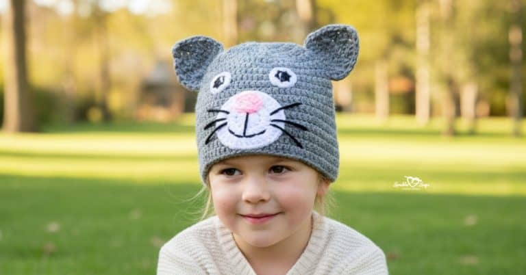Child wearing a gray crochet cat hat in a bright sunny park with green grass and softly blurred trees in the background
