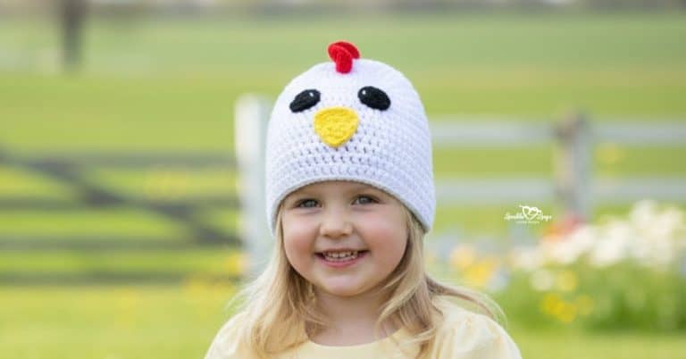 Child wearing a white crochet chicken hat in a sunny farm field with flowers and a fence in the background