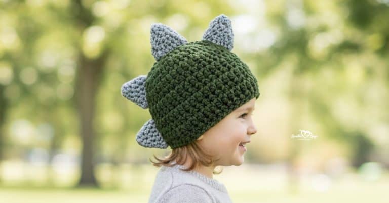 Side view of a child wearing a dark green crochet dinosaur hat with gray spikes in a bright sunny park with softly blurred trees in the background