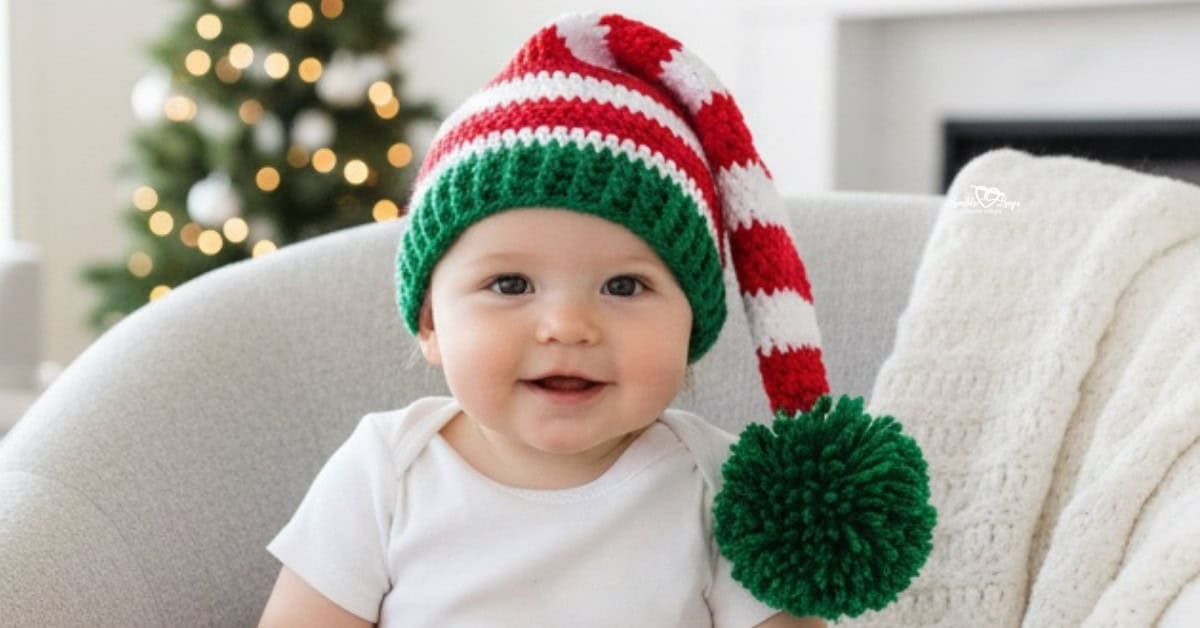 Baby wearing a red, white, and green striped crochet elf hat with a long tail and green pom-pom, sitting in a cozy chair with a Christmas tree in the background.