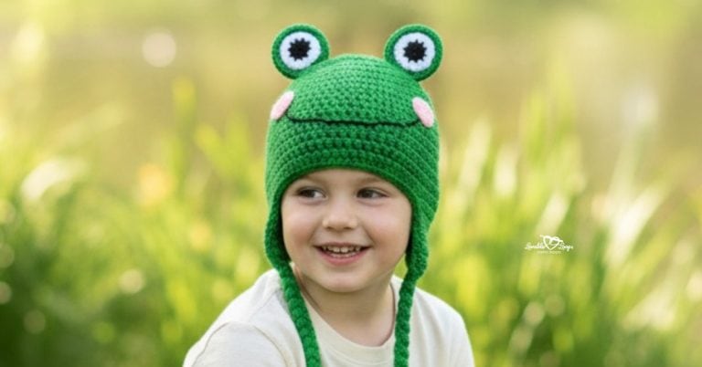 Child wearing a green crochet frog hat in a bright grassy pondside scene with soft greenery in the background