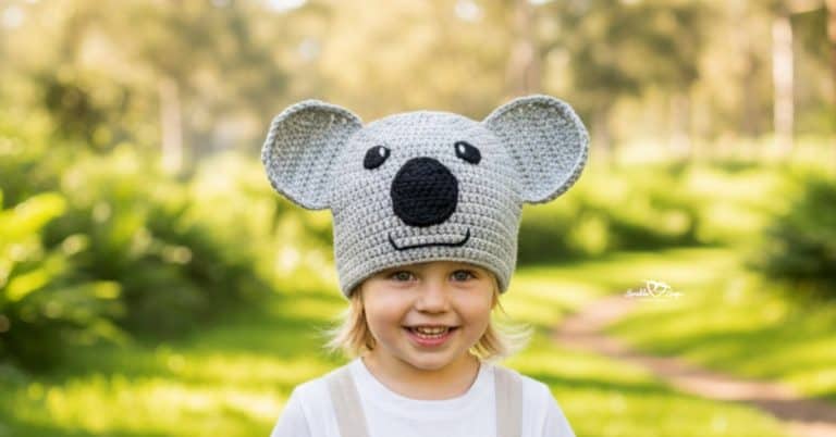 Child wearing a gray crochet koala bear hat on a bright green nature trail with soft trees in the background