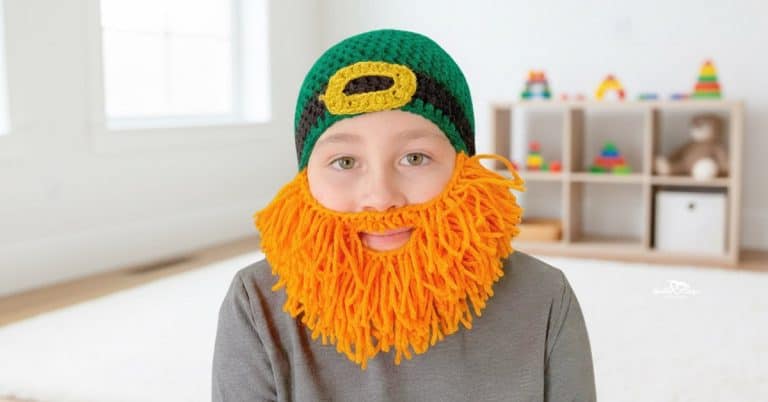 Boy wearing a green crochet leprechaun hat with a black-and-gold buckle and a bright orange yarn beard in a bright modern boys’ room.