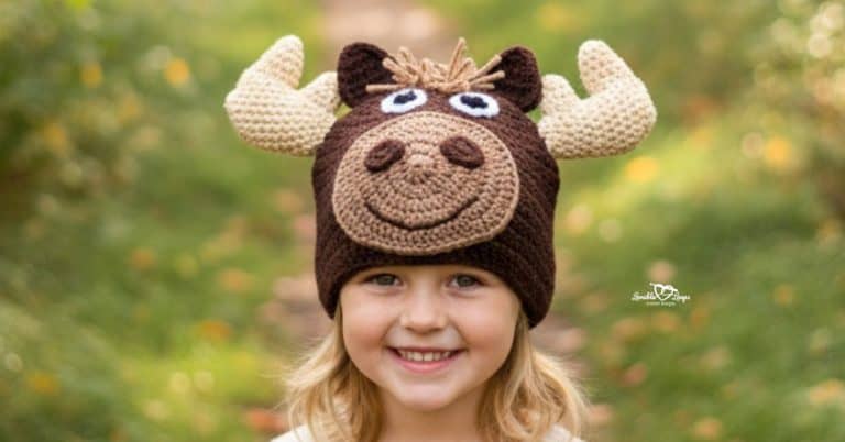 Child wearing a brown crochet moose hat with cream antlers on a green woodland trail with soft fall foliage in the background