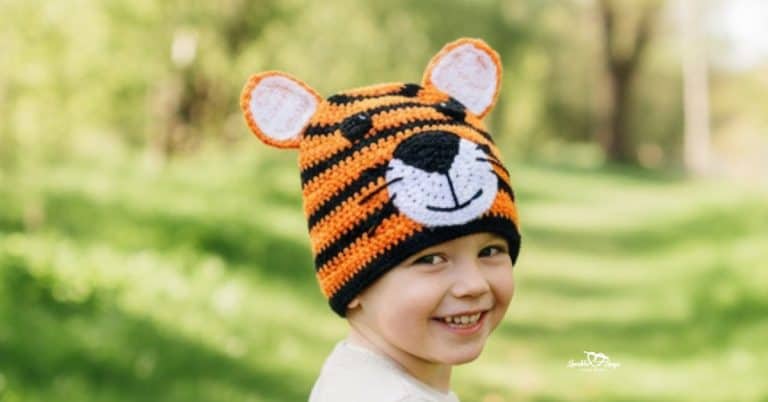 Child wearing an orange and black crochet tiger hat on a sunny green nature trail with trees in the background