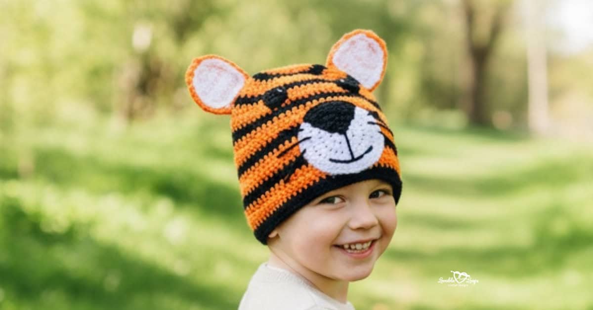 Child wearing an orange and black crochet tiger hat on a sunny green nature trail with trees in the background