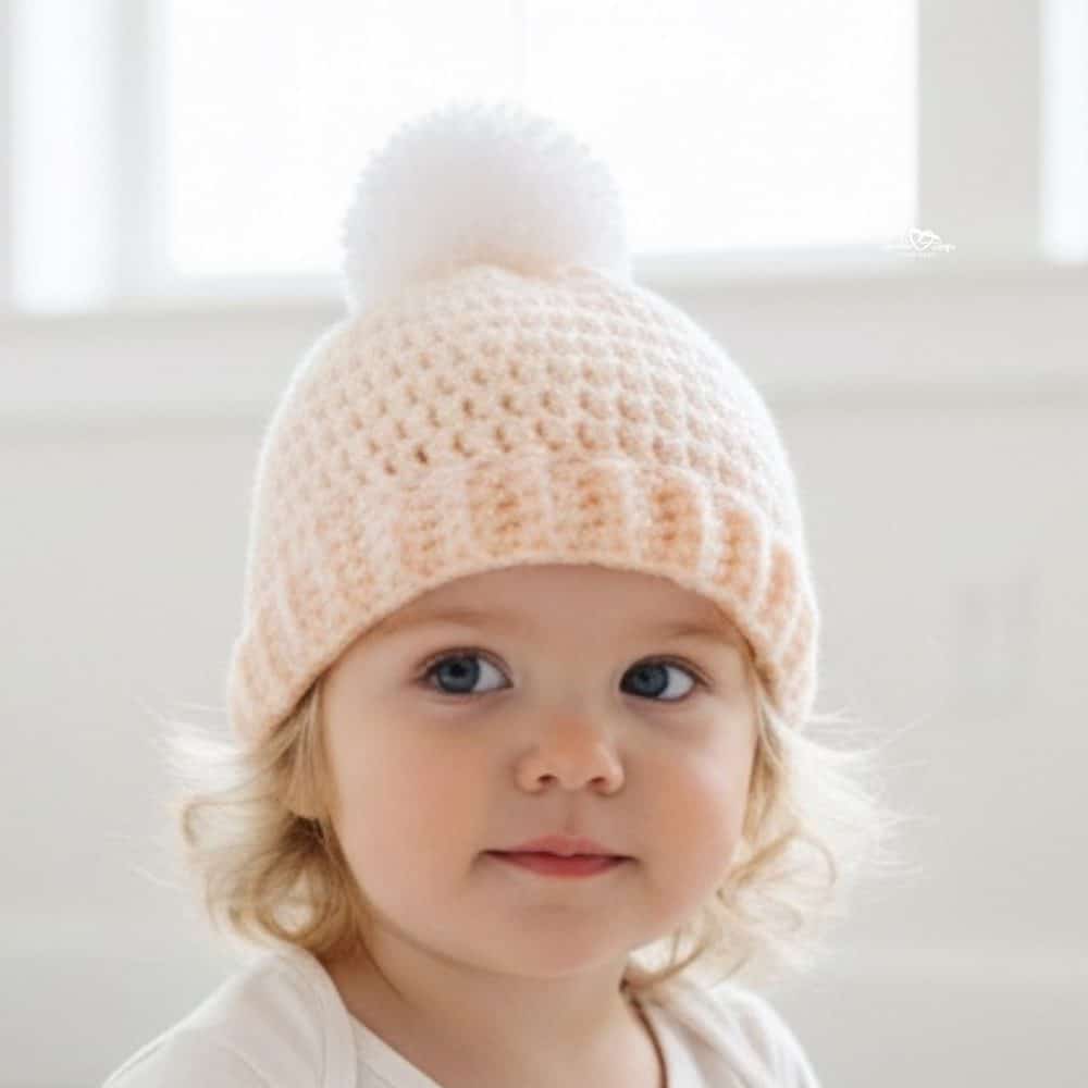Toddler wearing a soft peach crochet hat with a folded brim and fluffy white pom-pom against a light neutral background.