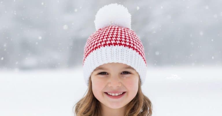 Girl wearing a red and white houndstooth crochet beanie with a white pom-pom standing outside in falling snow