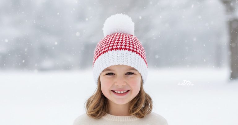 Girl wearing a red and white houndstooth crochet beanie with a white pom-pom standing outside in falling snow