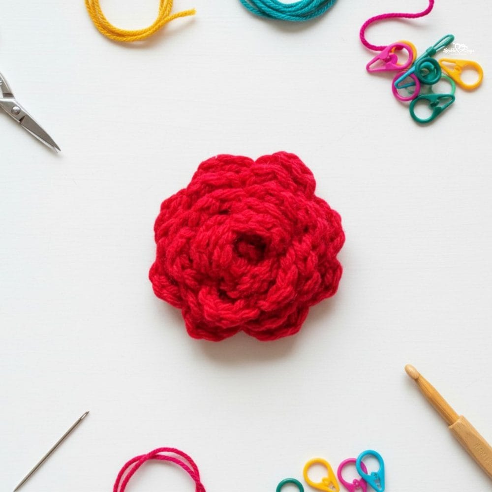 Red crochet rose applique on a white background with scissors, a crochet hook, yarn, a yarn needle, and colorful stitch markers nearby.