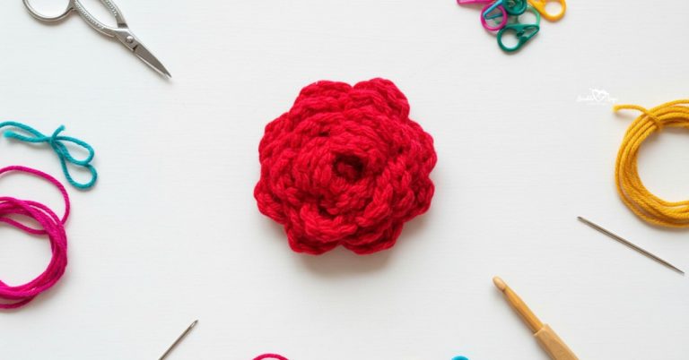 Crochet rose applique in bright red yarn centered on a white desktop with scissors, a crochet hook, yarn, a yarn needle, and colorful stitch markers around the edges.