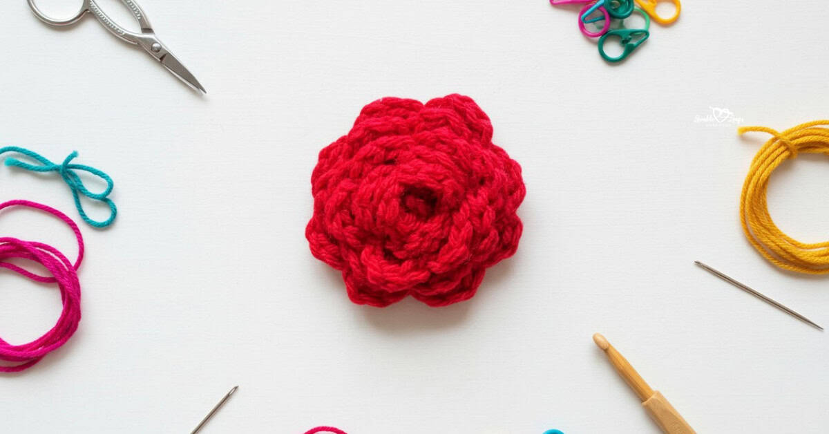 Crochet rose applique in bright red yarn centered on a white desktop with scissors, a crochet hook, yarn, a yarn needle, and colorful stitch markers around the edges.
