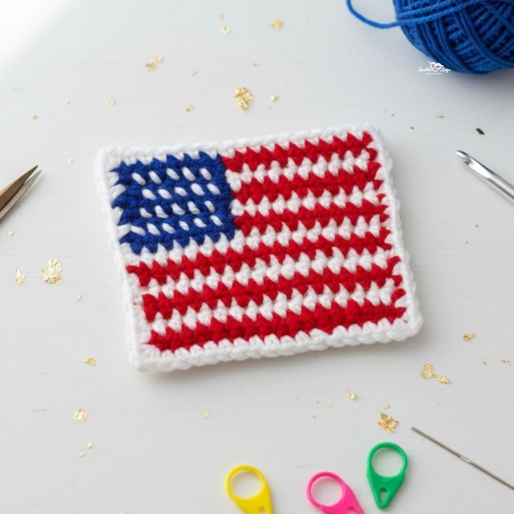 Crochet American flag applique in red, white, and blue yarn on a white desktop with scissors, a crochet hook, gold confetti, a yarn needle, and colorful stitch markers nearby.