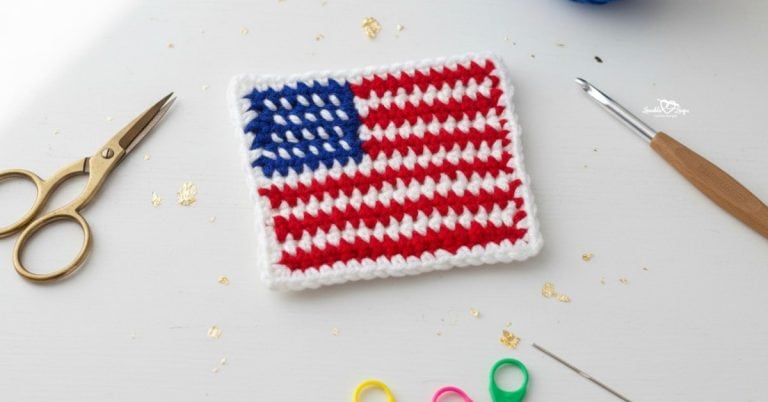 Crochet American flag applique centered on a white desktop with scissors, a crochet hook, gold confetti, a yarn needle, blue yarn, and colorful stitch markers around the edges.