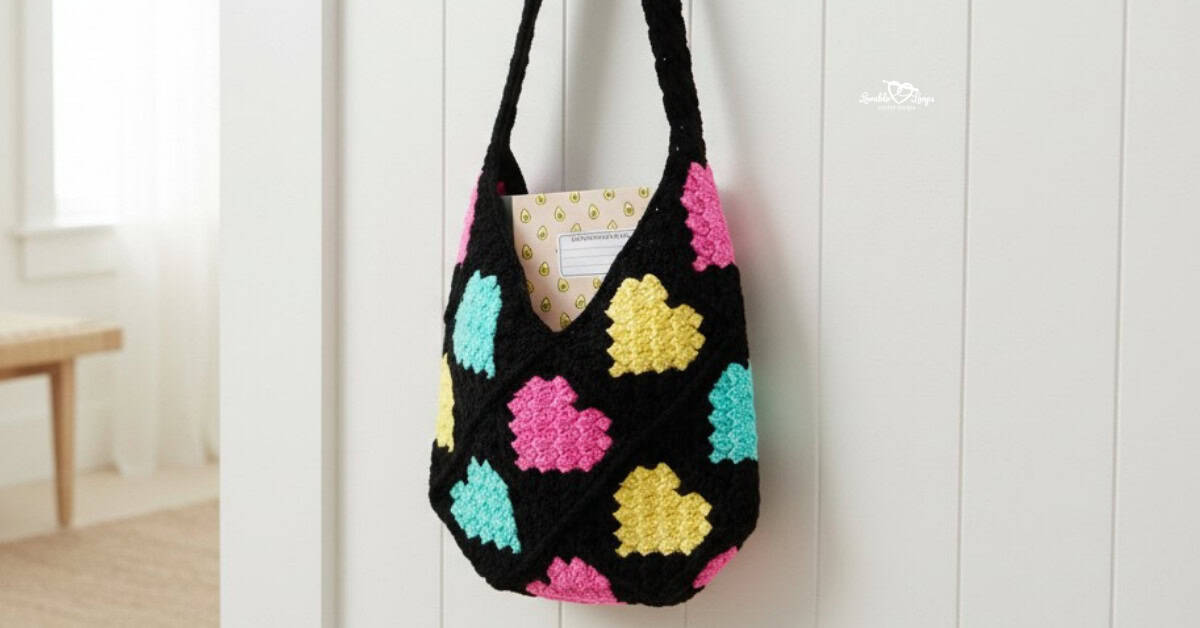 Black crochet tote bag with bright pink, yellow, and aqua heart motifs hanging on a light paneled wall with a notebook tucked inside.