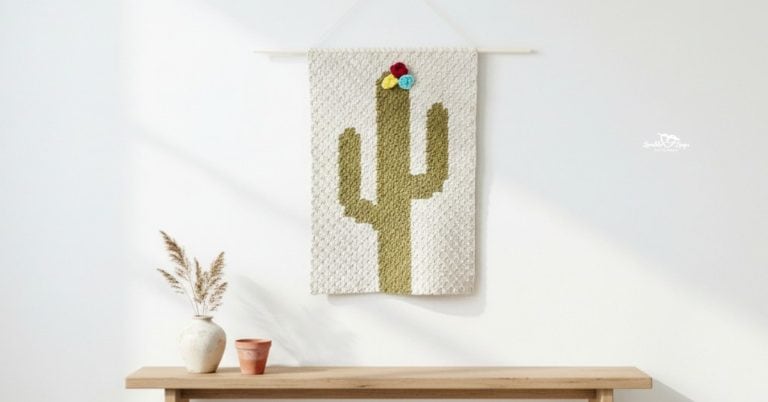 Crochet cactus wall hanging on a bright white wall above a light wood bench, styled with a small vase of dried stems and a terra-cotta pot.