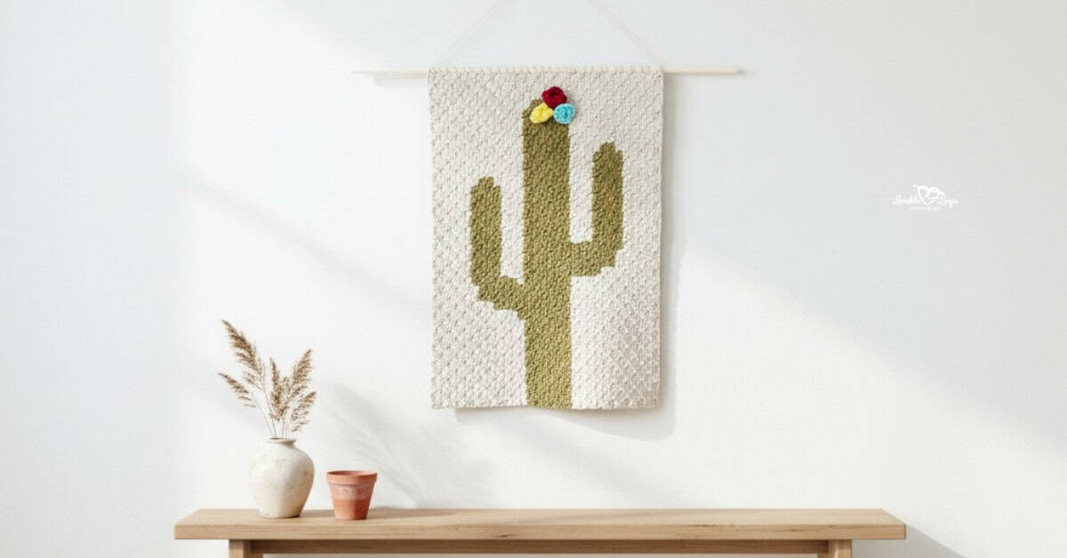 Crochet cactus wall hanging on a bright white wall above a light wood bench, styled with a small vase of dried stems and a terra-cotta pot.