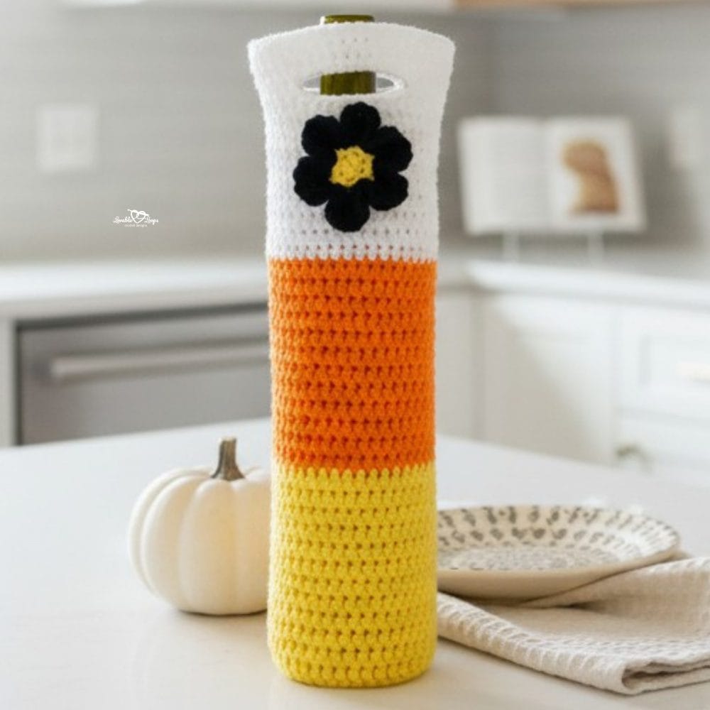 Candy corn crochet wine bottle bag with a black flower detail displayed on a bright kitchen island with soft fall decor and a modern white kitchen background.