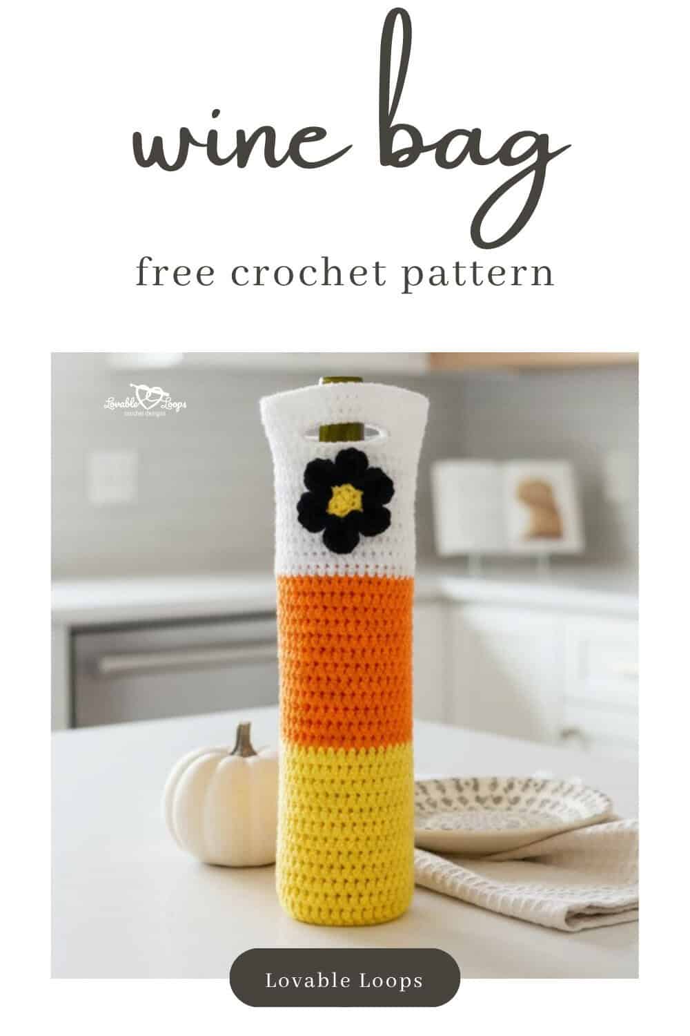 White, orange, and yellow crochet wine bottle bag with a black flower detail standing on a bright kitchen island beside a small white pumpkin, neutral dish, and cream napkin.