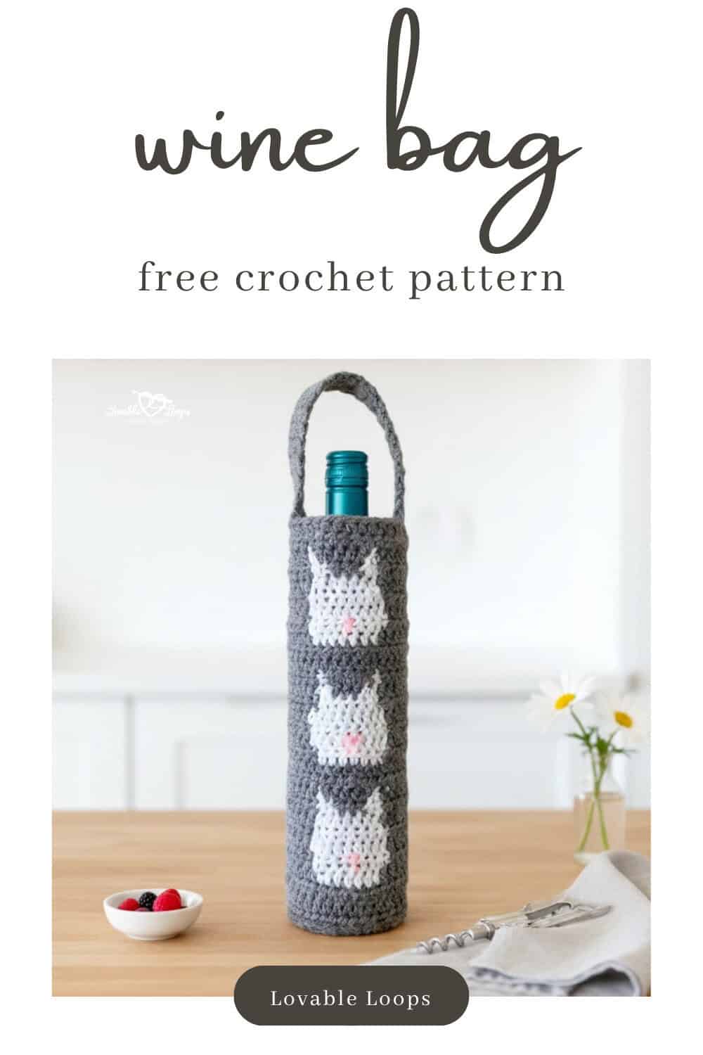 Gray crochet wine bottle bag with three white cat faces displayed on a light wood kitchen island with a small vase of daisies, a cream napkin, and a corkscrew.