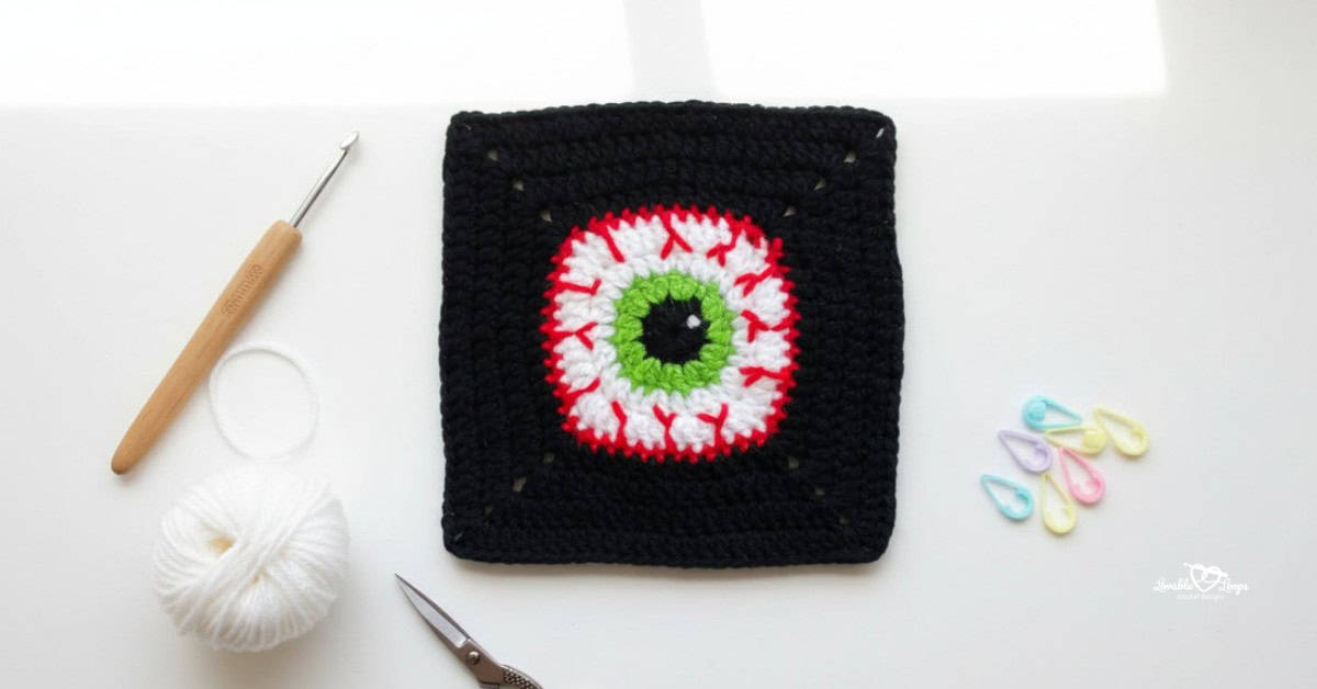 Overhead view of a black crochet granny square with a spooky eyeball design in white, red, green, and black, styled on a white desk with a crochet hook, white yarn, scissors, and pastel stitch markers.