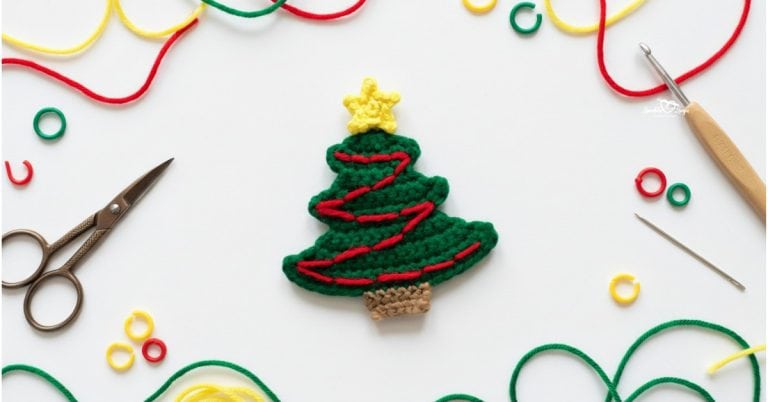 Crochet Christmas tree applique centered on a white desktop with colorful yarn, scissors, a crochet hook, a yarn needle, and stitch markers around the edges.