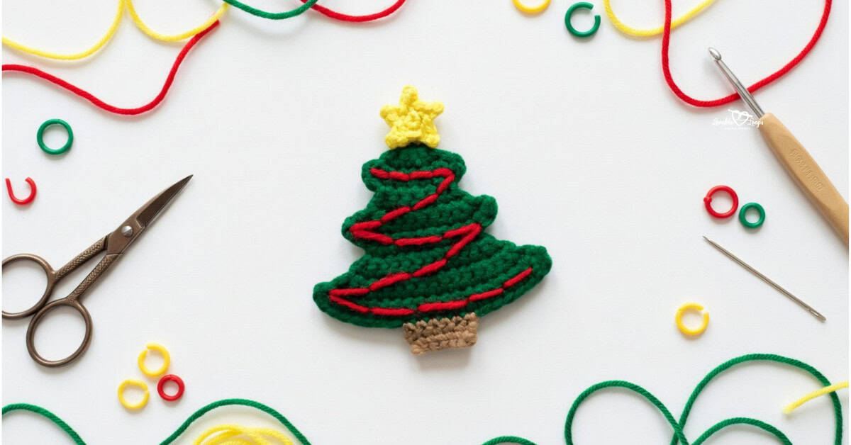 Crochet Christmas tree applique centered on a white desktop with colorful yarn, scissors, a crochet hook, a yarn needle, and stitch markers around the edges.