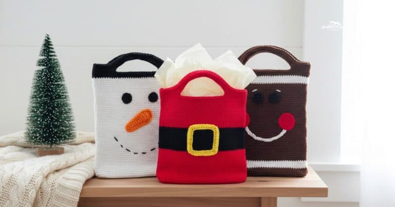Three crochet Christmas treat bags on a light wood bench, including a snowman bag, Santa suit bag, and gingerbread face bag, staged with tissue paper, a small bottlebrush tree, and a cream knit blanket.