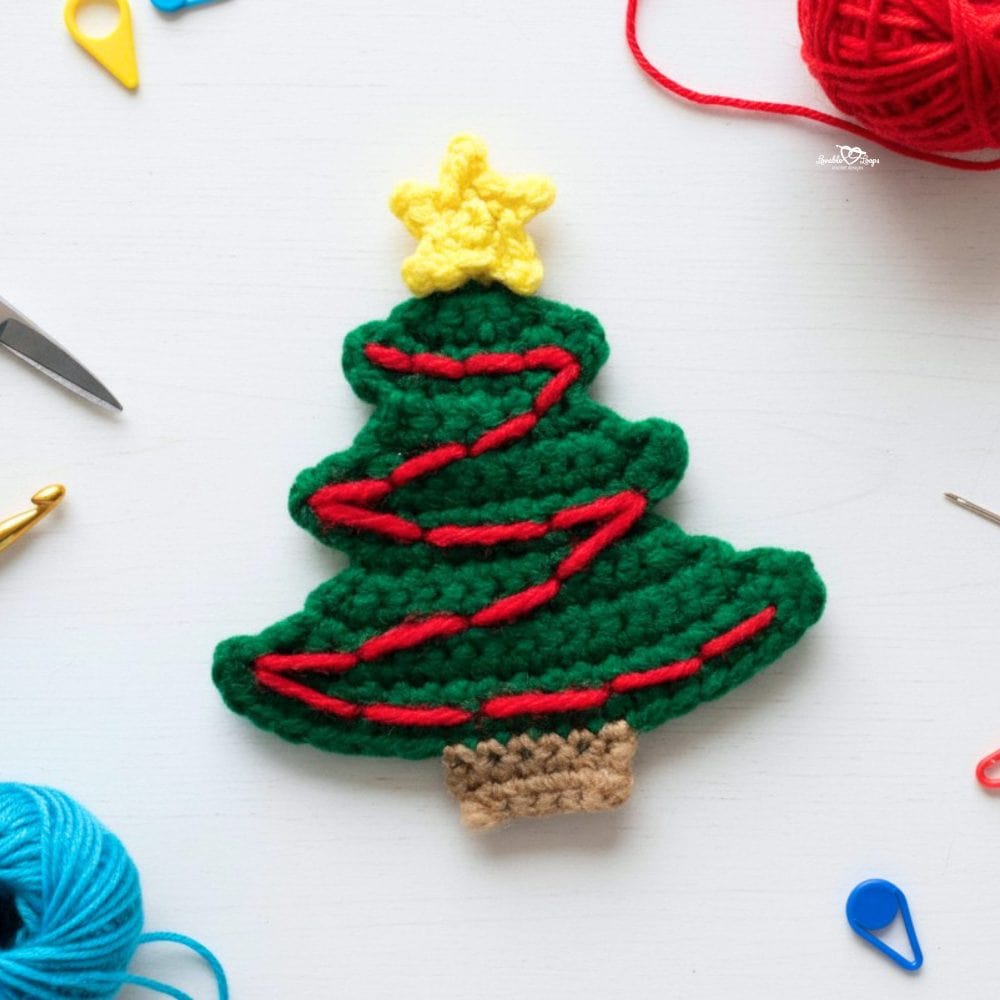 Crochet Christmas tree applique in green yarn with a red garland, yellow star, and brown trunk on a white background with yarn and crochet tools nearby.
