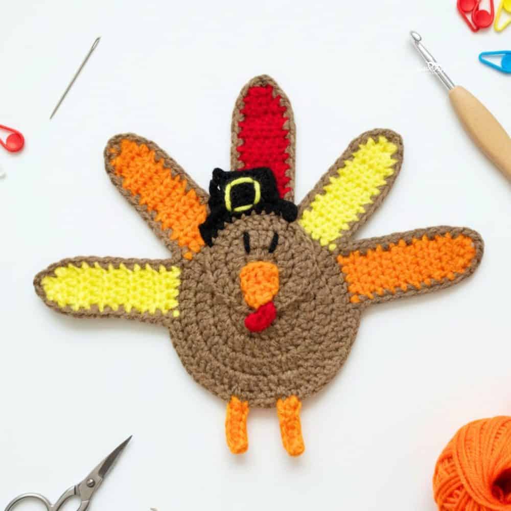 Crochet turkey applique in brown, orange, yellow, red, and black yarn with a pilgrim hat on a white desktop with scissors, a crochet hook, yarn, and stitch markers nearby.